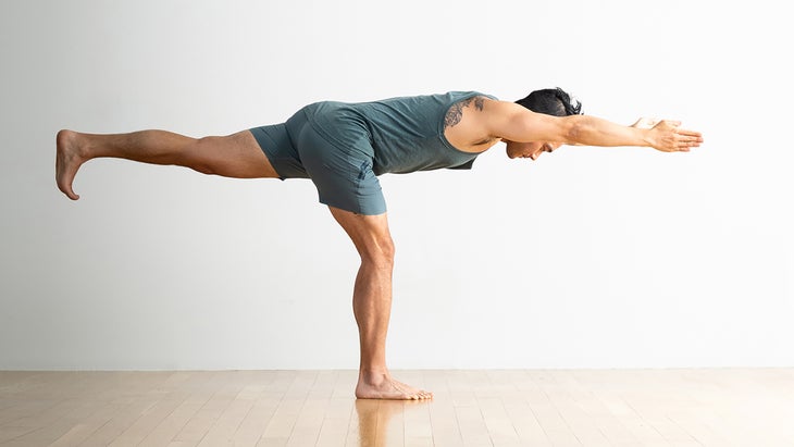 Person in Warrior III Pose. He is wearing teal blue shorts and a sleeveless top. He is standing on a wood-plank floor with a plain white wall behind him.