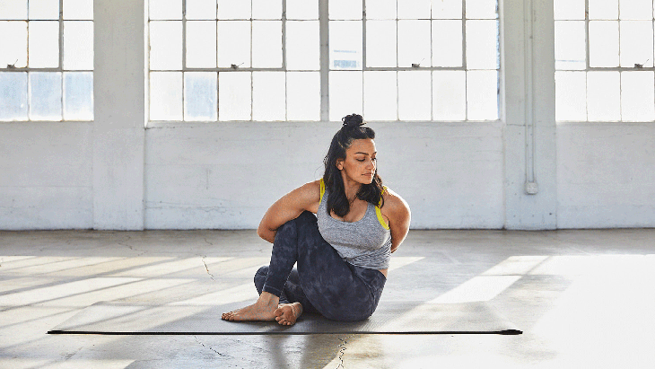 Woman sitting on a yoga mat with her knees bent twisting to the side and looking over her shoulder in a Lotus Pose modification