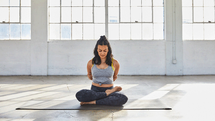 Woman sitting on her yoga mat with her knees bent and her arms behind her in a Half Bound Lotus Pose modification