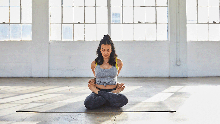 Woman sitting on a yoga mat with her legs crossed in Lotus Pose and her arms behind her back