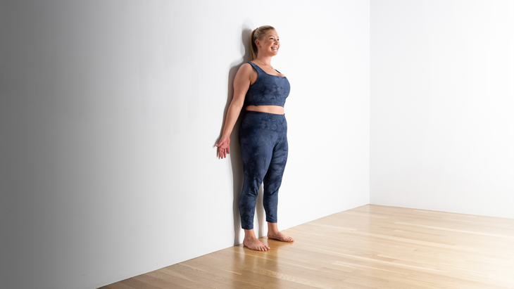 A blonde woman stands against a wall in Mountain Pose.