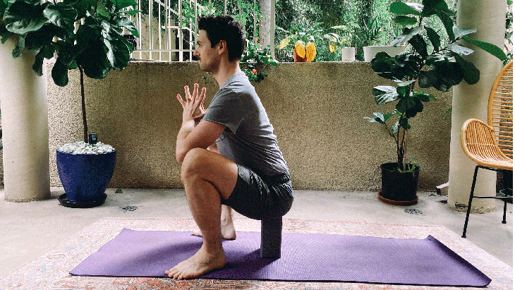 Man learning how to practice Malasana, or yoga squat, with a block beneath his seat for support and his hands at his chest in prayer