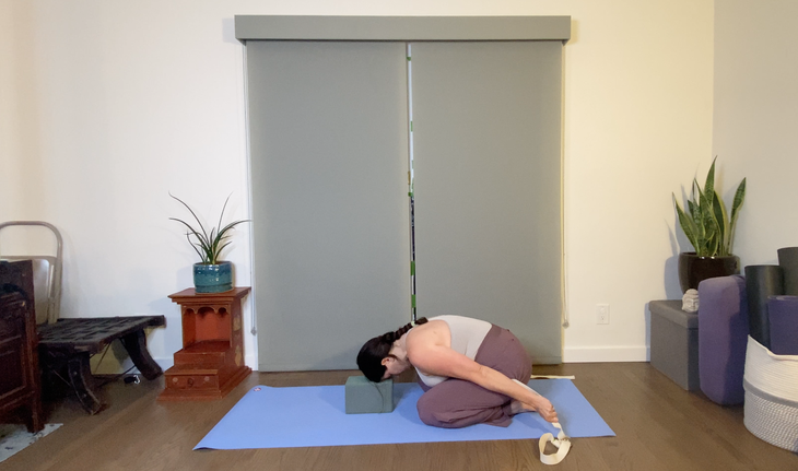 A dark-haired woman practices Balasana with her head on a yoga block.