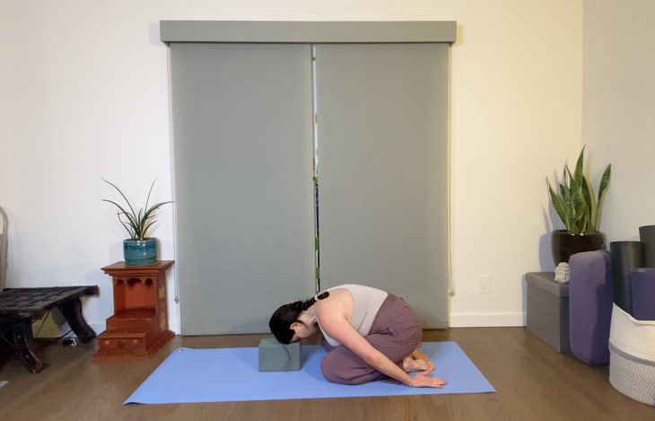 Woman in purple pants practices Child's Pose with her head on a block