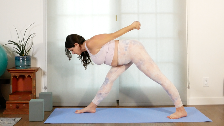 A woman with dark hair practices Parsvottanasana with an arm variation. She is wearing pastel colored yoga tights and a top. There is a window behind her that is covered with sheer curtain blinds. In the background there are plants, a large green ball, and a small wooden table.
