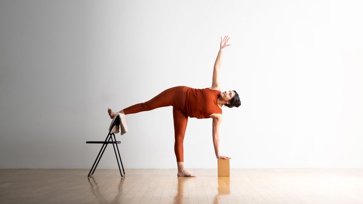 Noemi Nunez practices Half Moon Pose (Ardha Chandrasana) with her foot on a metal folding chair. She has dark hair and is wearing copper colored clothes. She is on a light wood floor with a white wall in the background.