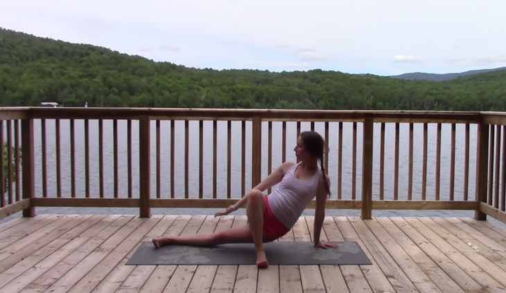 Yoga With Kassandra seated cross-legged outside on a mat practicing a low lunge with a twist