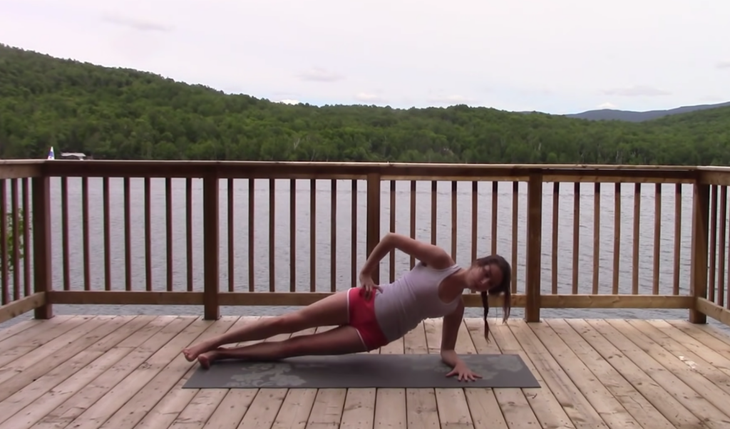 Yoga With Kassandra practices a forearm side plank on a mat outside with her right hand on her right hip