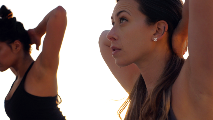 Women on a rooftop practicing yoga with their hands behind their heads and their fingers interlaced.